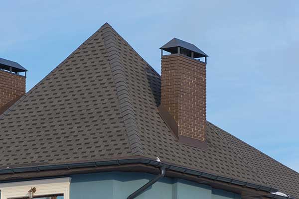 Close up of a dark brown shingle roof with two brick chimneys with brown flashing and caps
